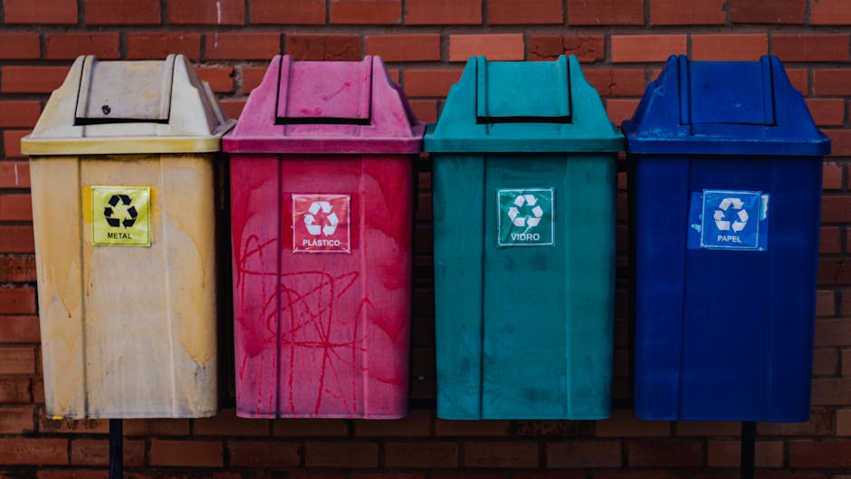 Four colour-coded recycling bins aligned against a red brick wall, each with a distinct label indicating their contents for household waste sorting. The first bin on the left is pale yellow with a bla