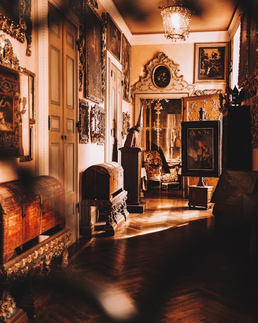 An interior view of an ornate room with antique furniture and decorative artwork, illuminated by warm lighting from a chandelier. The space features wooden flooring and walls adorned with framed paint